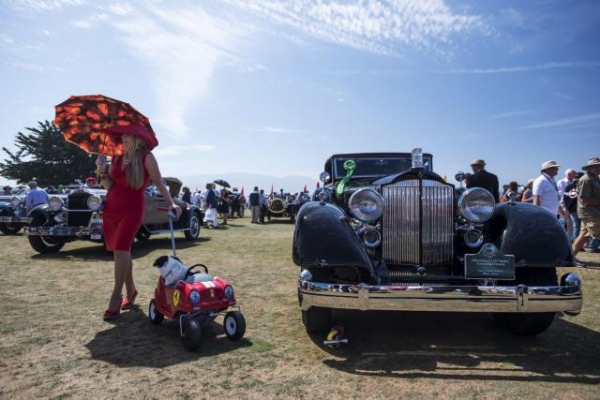 Una azafata posa sosteniendo un paraguas y tirando de un carrito de Ferrari en el que va subido su perro para una fotografía al lado de un Packard 1107 Victoria convertible de 1934, a la derecha, durante el Pebble Beach Concours d’Elegance en Pebble Beach, California (Estados Unidos).